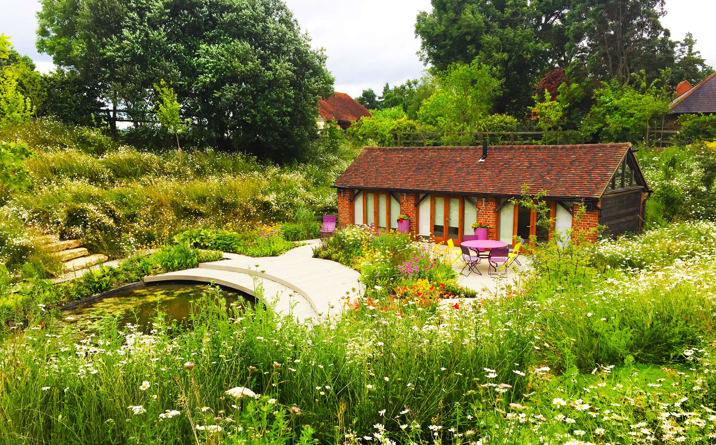 Red brickwork building to right hand side of image, bright purple contemporary seating looking out towards lower wildlife pond. Surrounded by light wooden decking, with beautiful naturalistic, pollinator friendly planting beds surrounding. Large mature trees in the background. Beautiful meadow planting in the foreground creating garden landscapes rich in biodiversity. Designed by garden designer Ann-Marie Powell Gardens based in Hampshire.